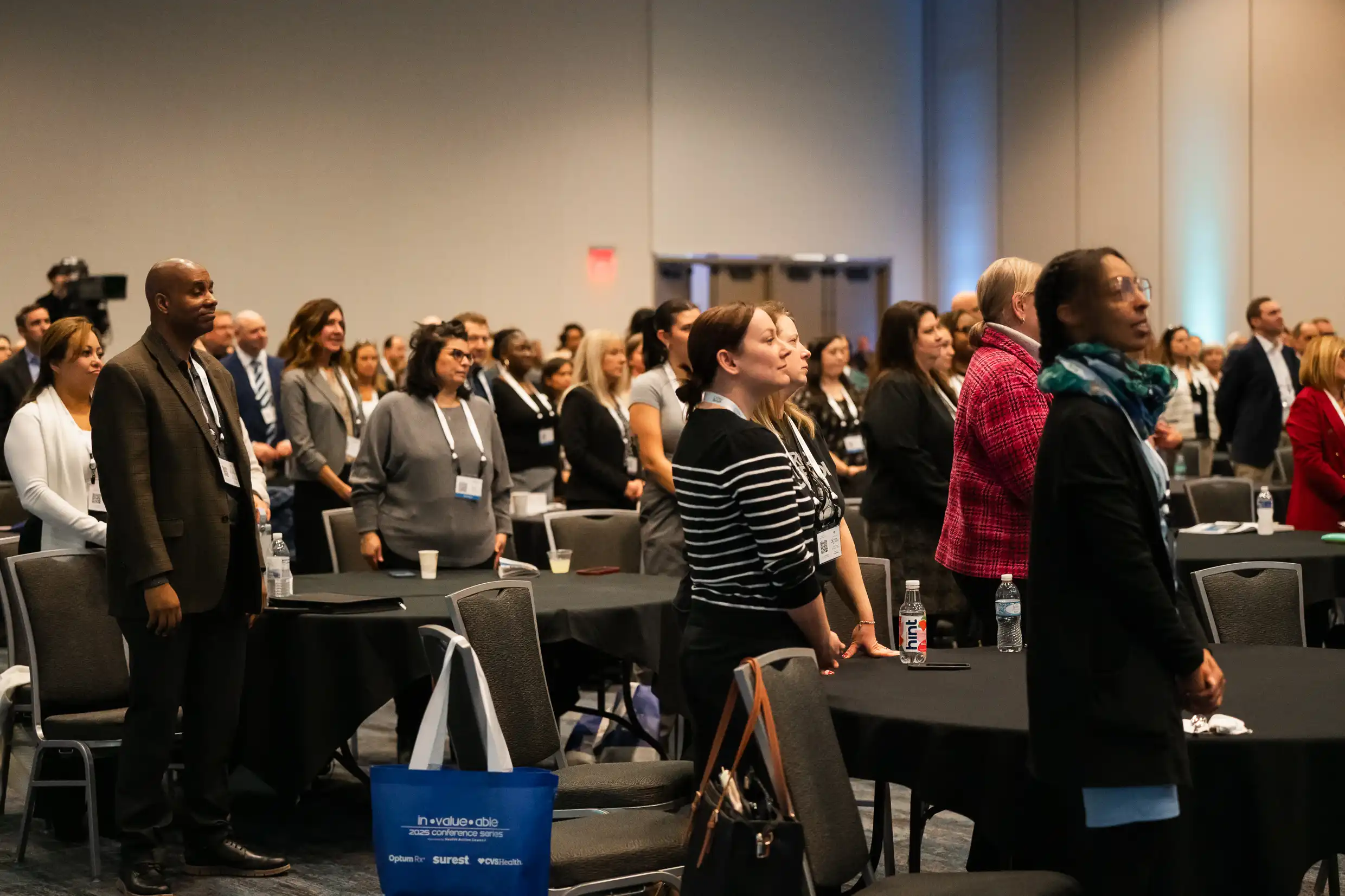 Audience Members Standing Diverse audience standing during In-Value-Able 2025 keynote session in large event hall, professional attendees with name badges and branded tote bags.