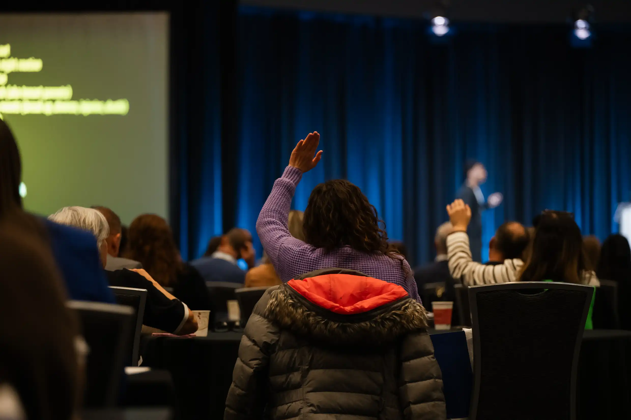 Audience member with raised arm Audience member raising hand during In-Value-Able 2025 conference Q&A session with speaker on stage and presentation screen in background.