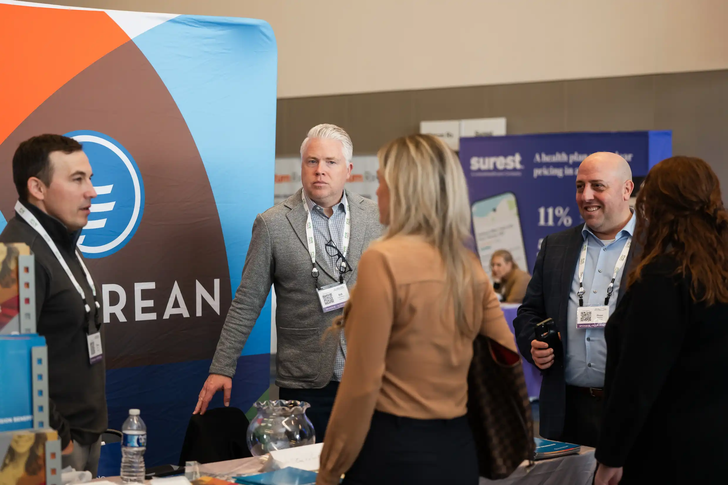 Showcase your Products A group of professionals engage in conversation at a In-Value-Able 2025 booth for Empyrean, a benefits administration company. Two men stand behind the booth, which features a colorful branded backdrop, while two women stand in front speaking with them. Other booths and attendees are visible in the background, including one for Surest, another health-related company.