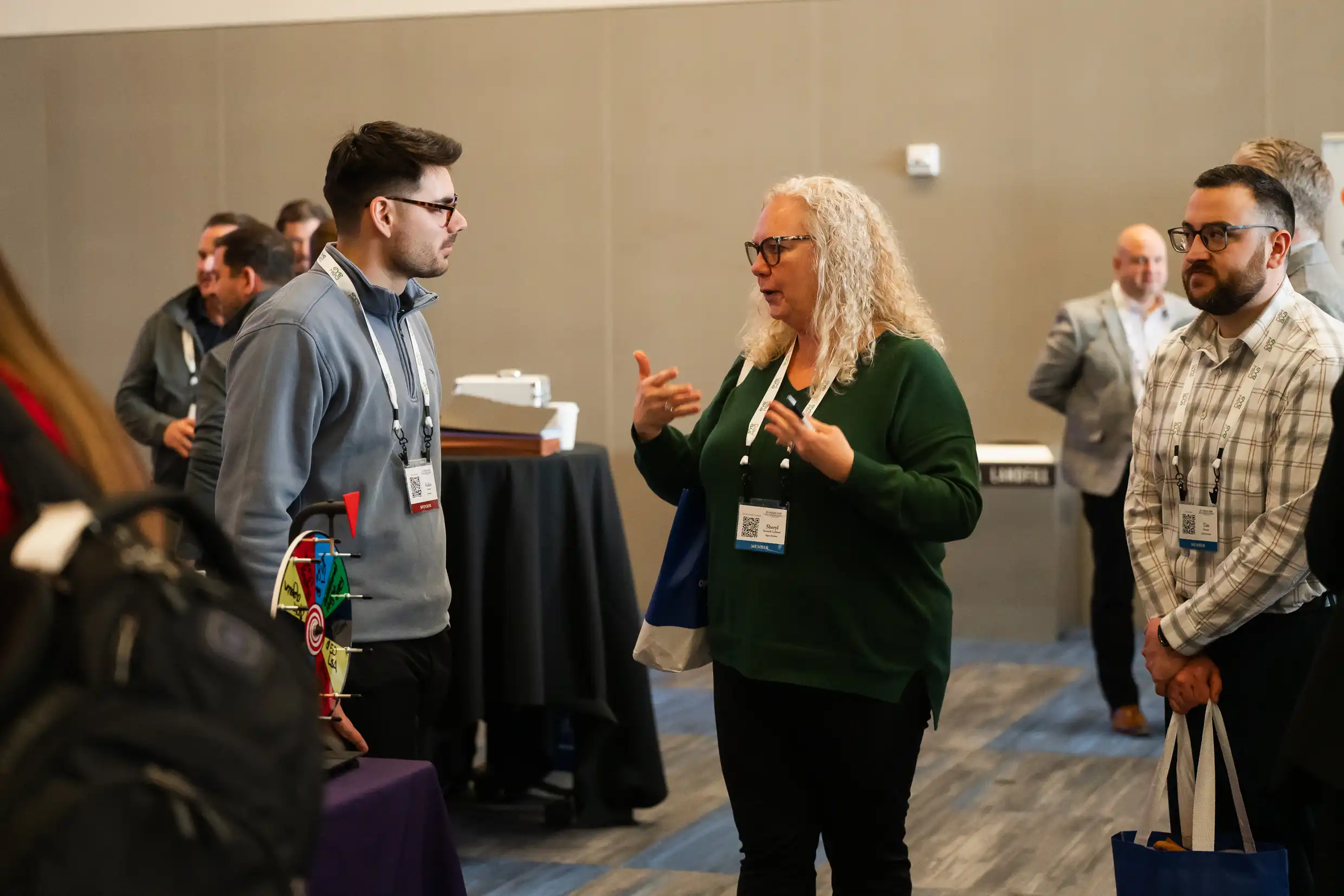 Members talking during break A group of conference attendees engage in conversation during a break. A woman with curly blonde hair and glasses, wearing a green sweater, speaks animatedly to a man in a gray pullover. Another man in a plaid shirt stands nearby holding a tote bag. Several people and high-top tables are visible in the background, along with a small prize wheel on a table in the foreground.