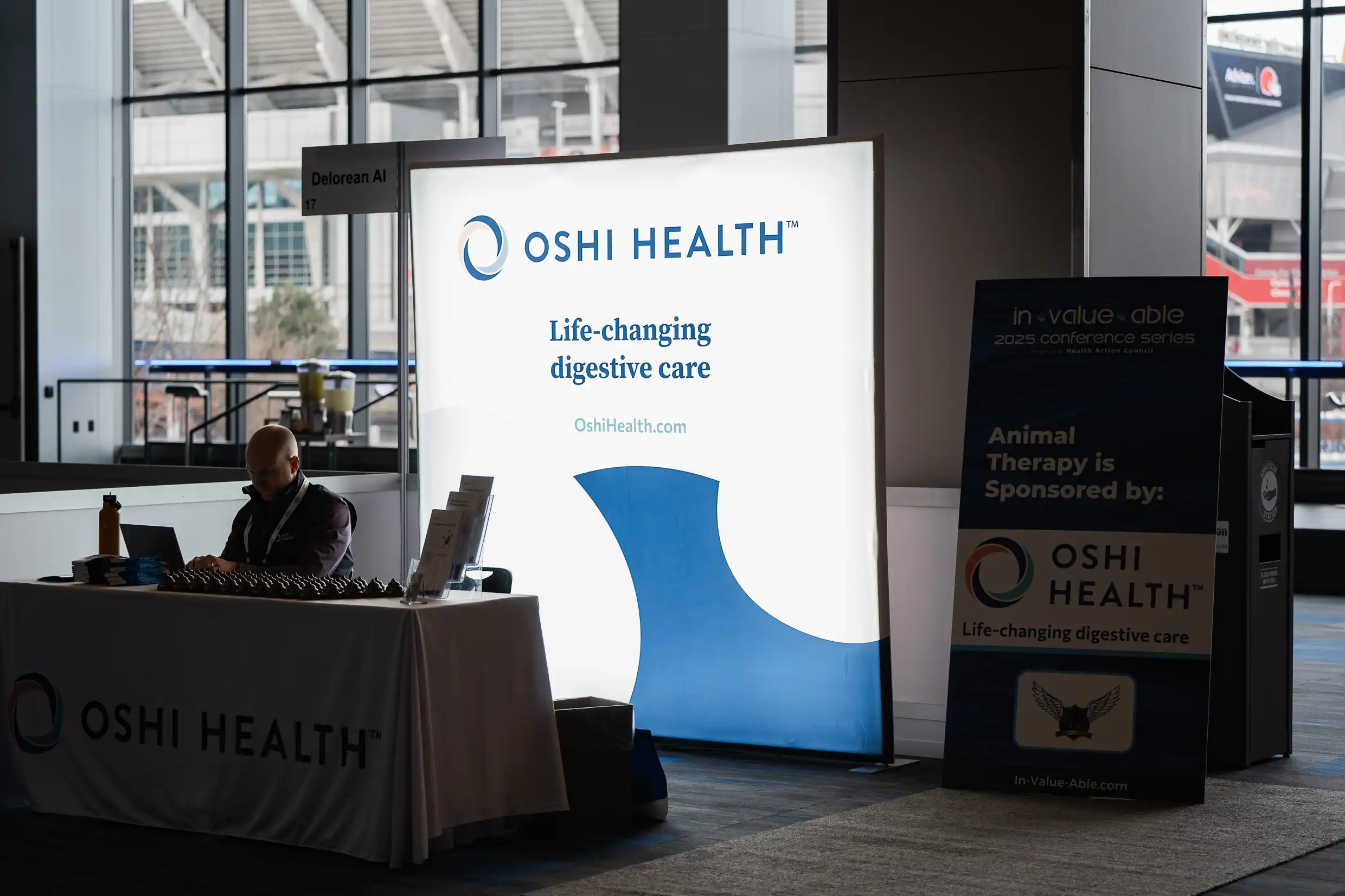 Oshi Health booth Branding A conference exhibitor booth for Oshi Health, featuring a large illuminated sign with the slogan "Life-changing digestive care" and the website OshiHealth.com. A person sits at the booth working on a laptop, with promotional materials and giveaways displayed on the table. A separate sign nearby indicates Oshi Health is sponsoring animal therapy for the 2025 conference series. The setting includes large windows showing an exterior view of a stadium or arena.