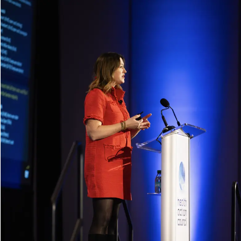 A female keynote speaker in a vibrant red dress delivers a presentation at the Health Action Council conference, standing at a professional white podium against a blue-lit stage.